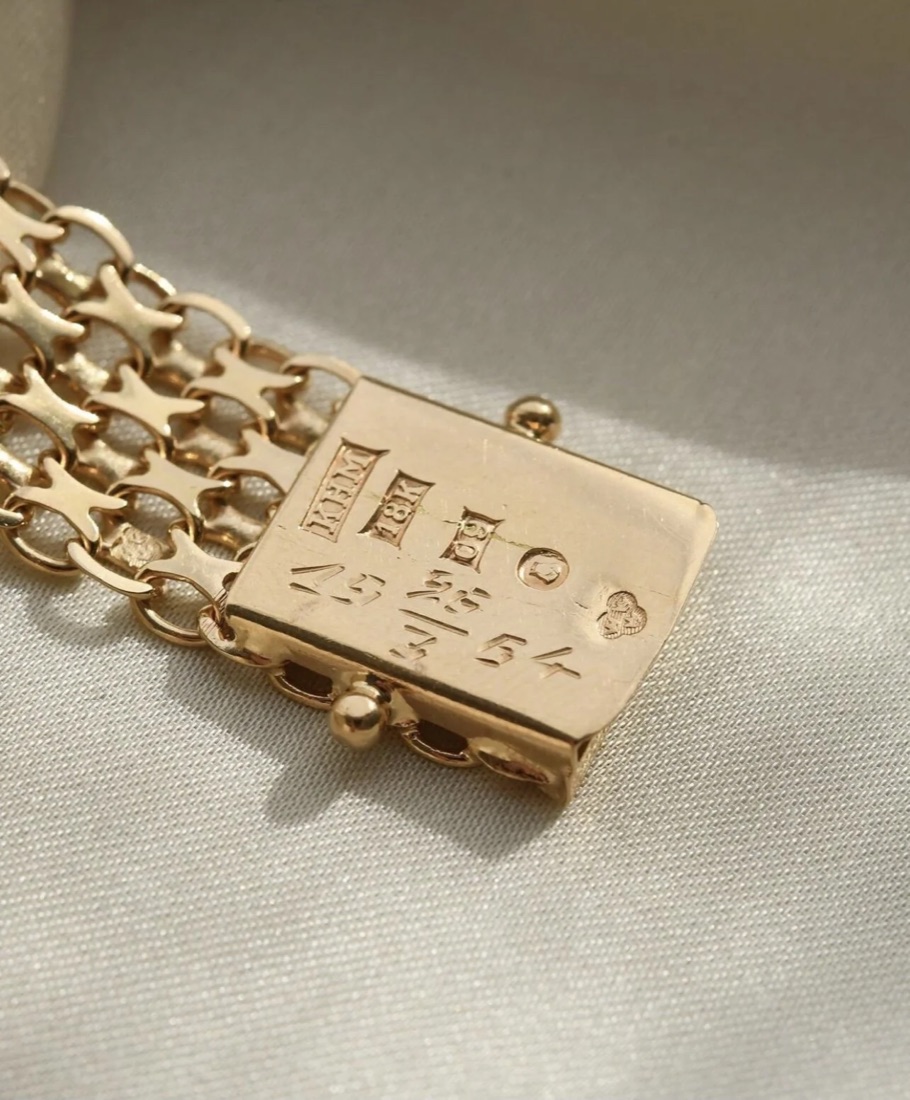 The clasp of a gold bracelet, photographed close up. A row of five punches is struck into it: KFM in a rectangle, 18K in a rectangle, a letter with a small numeral beside it, a round town mark, and three crowns arranged in a trefoil.